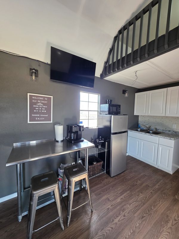 Modern kitchenette with stainless steel table, stools, and appliances in a cozy retreat space.