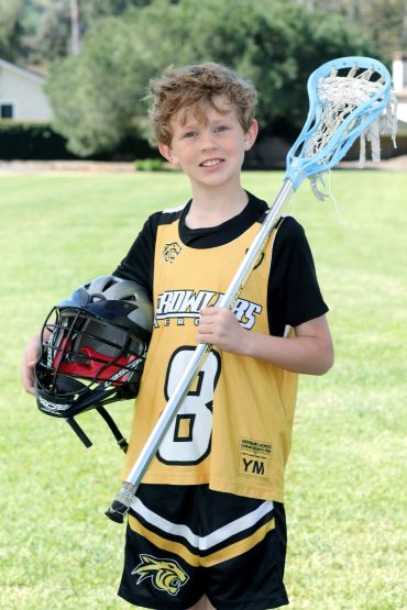 Young boy in lacrosse gear holding a helmet and lacrosse stick.