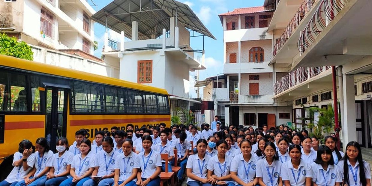 A large group of students in uniforms seated outdoors near a yellow school bus.
