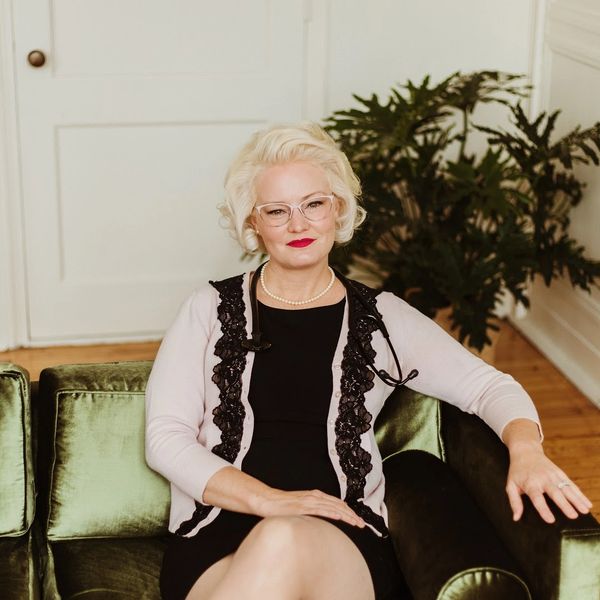 Confident woman in glasses and pearls sitting on a green velvet sofa.