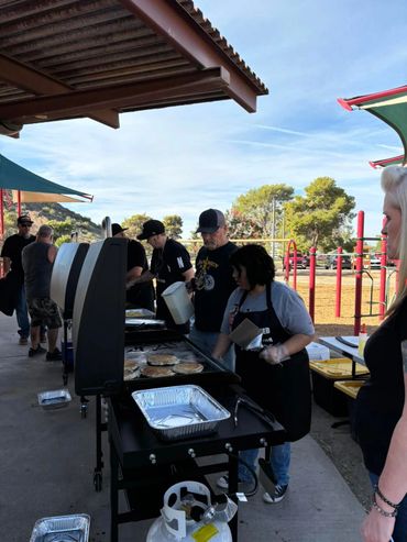People cooking pancakes on a large outdoor griddle under a covered area.