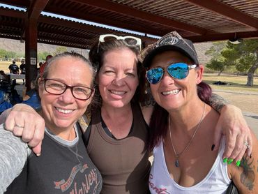 Three smiling women posing closely outdoors under a shelter.
