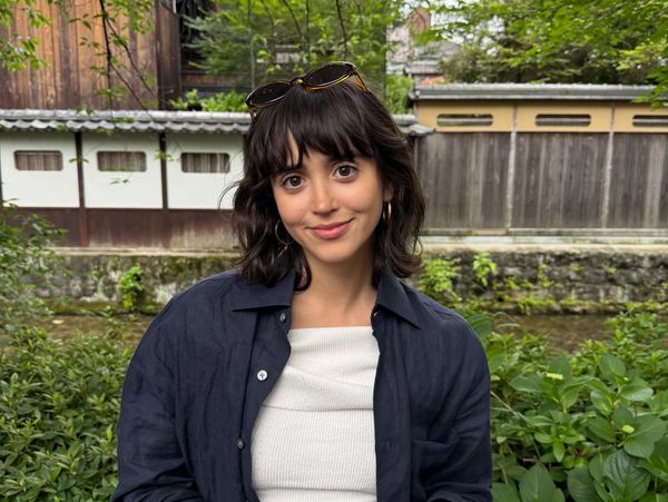 Young woman smiling outdoors with greenery and traditional buildings behind her.