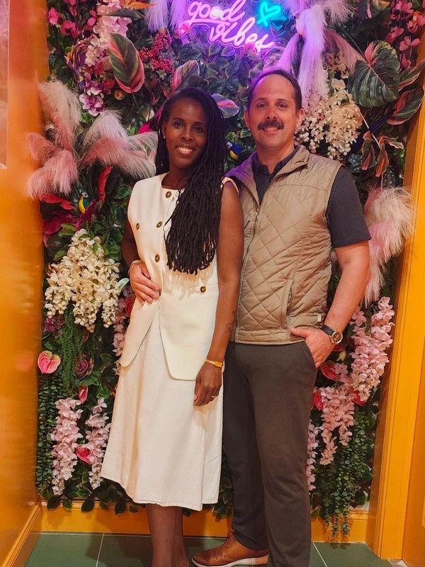 A couple poses happily in front of a vibrant floral wall with a neon 'good vibes' sign.