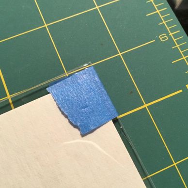 Blue painter's tape securing paper to a glass surface on a cutting mat.