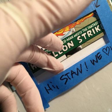 Gloved hand holding a comic book page with a taped note saying "Hi STAN! WE ♥".