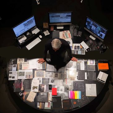 Person surrounded by documents and three computer screens in a dark room.