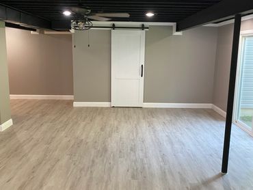 Painted black exposed basement ceiling with vinyl plank flooring and sliding single barn door.