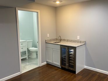 Basement wet bar with granite countertop, wine cooler, basement bathroom and vinyl flooring.