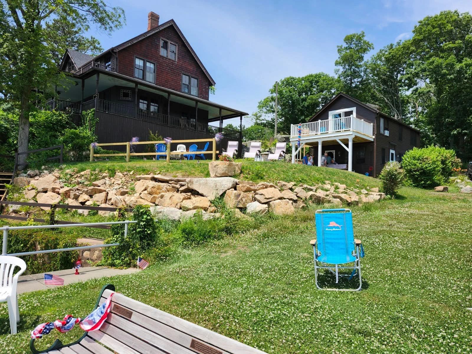 Two houses on a sunny grassy hill with chairs and a bench.