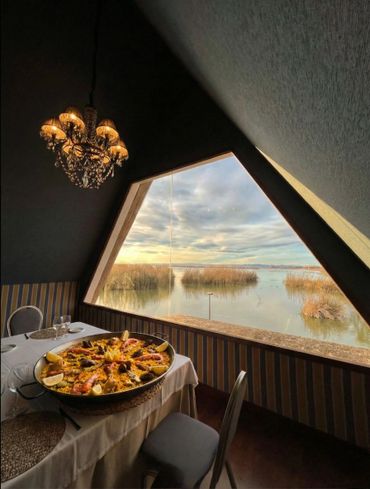 Elegant dining with seafood paella by a large geometric window overlooking a serene water view.