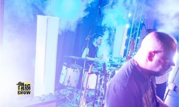 A drummer performs on stage surrounded by smoke and blue lighting.