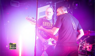 Two guitarists perform energetically on stage with vibrant purple lighting.