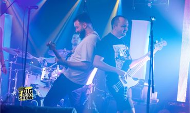 Two guitarists and a drummer energetically perform on stage with vibrant blue lighting.