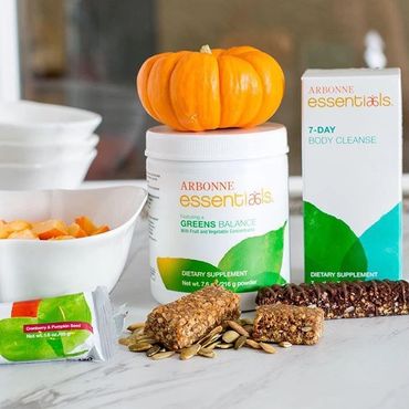 Arbonne Greens Balance Powder and 7-day cleanse Sachets with a protein bar on a table top.