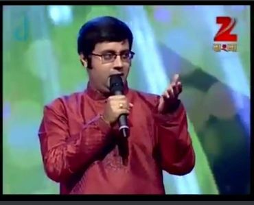 Man in red traditional attire speaking on microphone on stage.