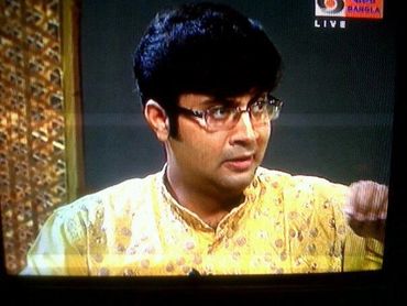 Man in glasses and yellow traditional attire speaking on live TV.