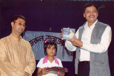 Three people at an event, one holding a CD and another holding flowers.