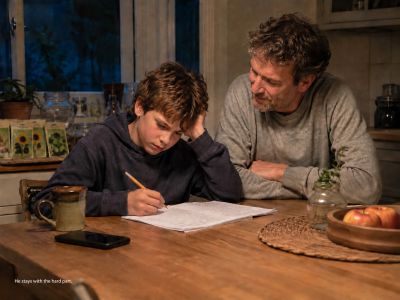 Boy writes at kitchen table; father watches, phone set aside.