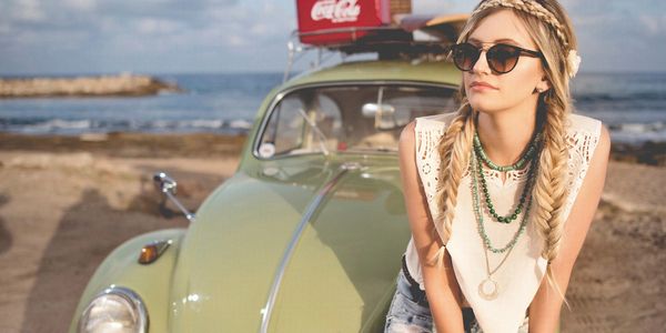 Blonde haired lady sitting on the front headlight of a  VW Beetle on the beach 