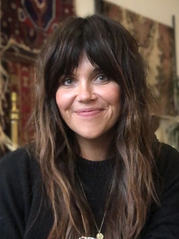 A woman with long brown hair and bangs smiles warmly at the camera.