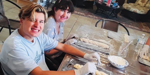 Two young people smiling while braiding dough at a kitchen table.