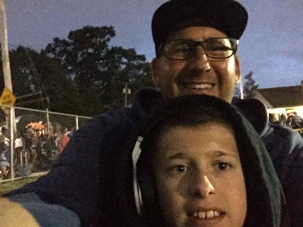 A man and a boy taking a selfie outdoors at dusk with a crowd in the background.
