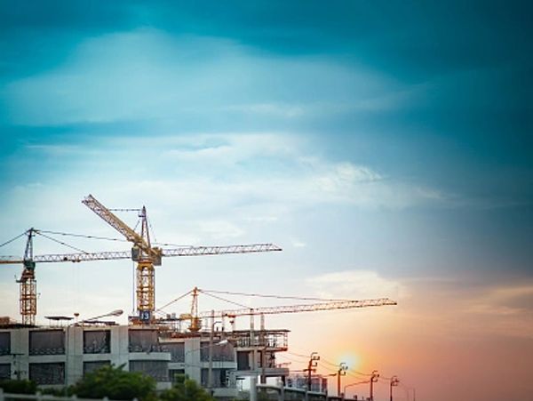 Large construction cranes stand tall against a dramatic sky at sunset, building a new structure.