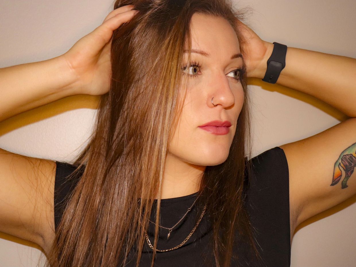 Woman with nose ring and tattoo, posing with hands in hair.