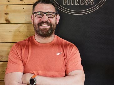 Smiling man in glasses and orange shirt standing in front of a fitness sign.