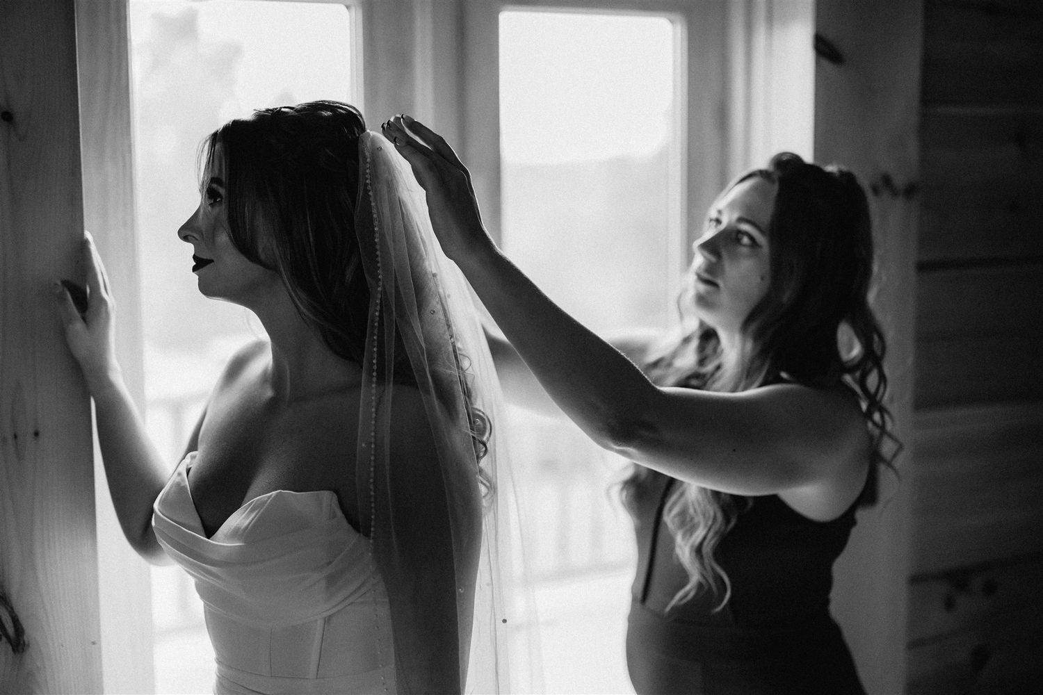 A bride stands by a window while another woman adjusts her veil in a black and white photo.