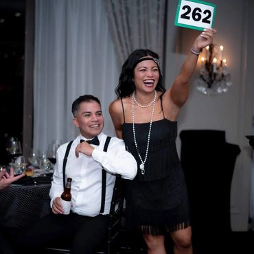 A joyful woman holds up an auction paddle with number 262, smiling next to a seated man with a beer bottle.