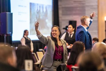 Smiling woman in a gray suit raises her hand at a conference or event.