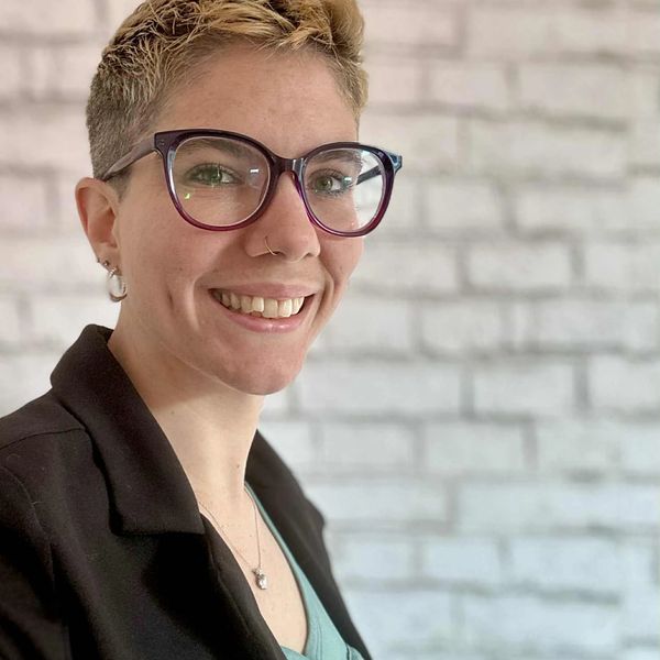 Smiling person with short hair and glasses in front of a white brick wall.