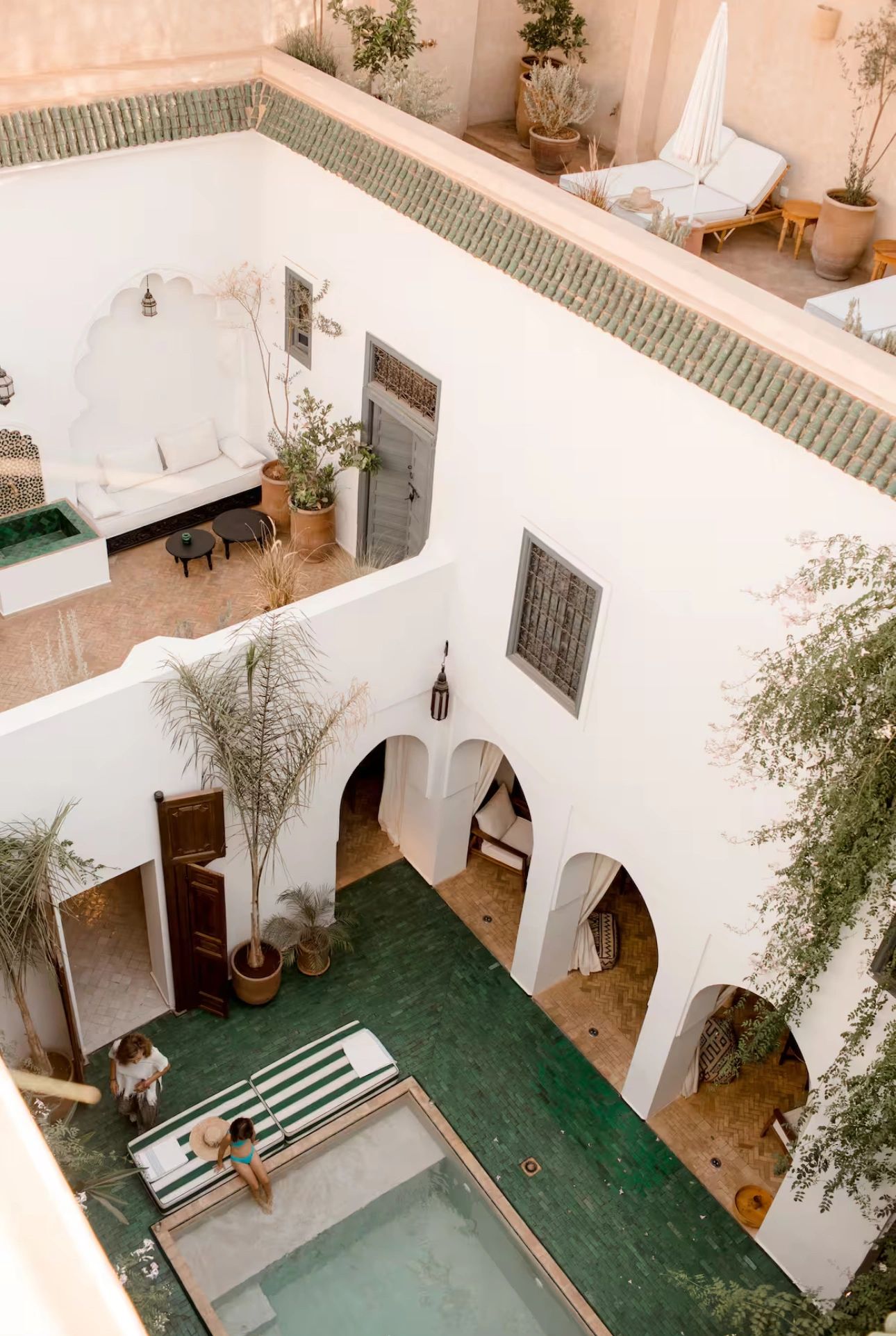 Sunlit Marrakech riad courtyard with a swimming pool and guests relaxing beside the water.