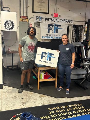 Two people holding a physical therapy sign in a gym setting.