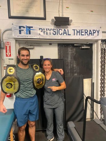 Two people smiling in a physical therapy clinic, one wearing championship belts.