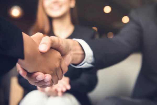Two people shaking hands in a business setting with a smiling woman in the background.