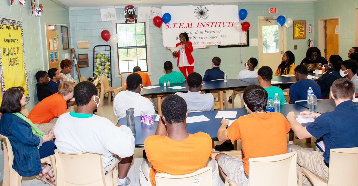A woman in a red suit speaks to a diverse group in a classroom setting.