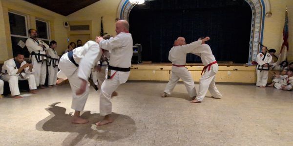 Two pairs of martial artists sparring in a dojo with an audience.