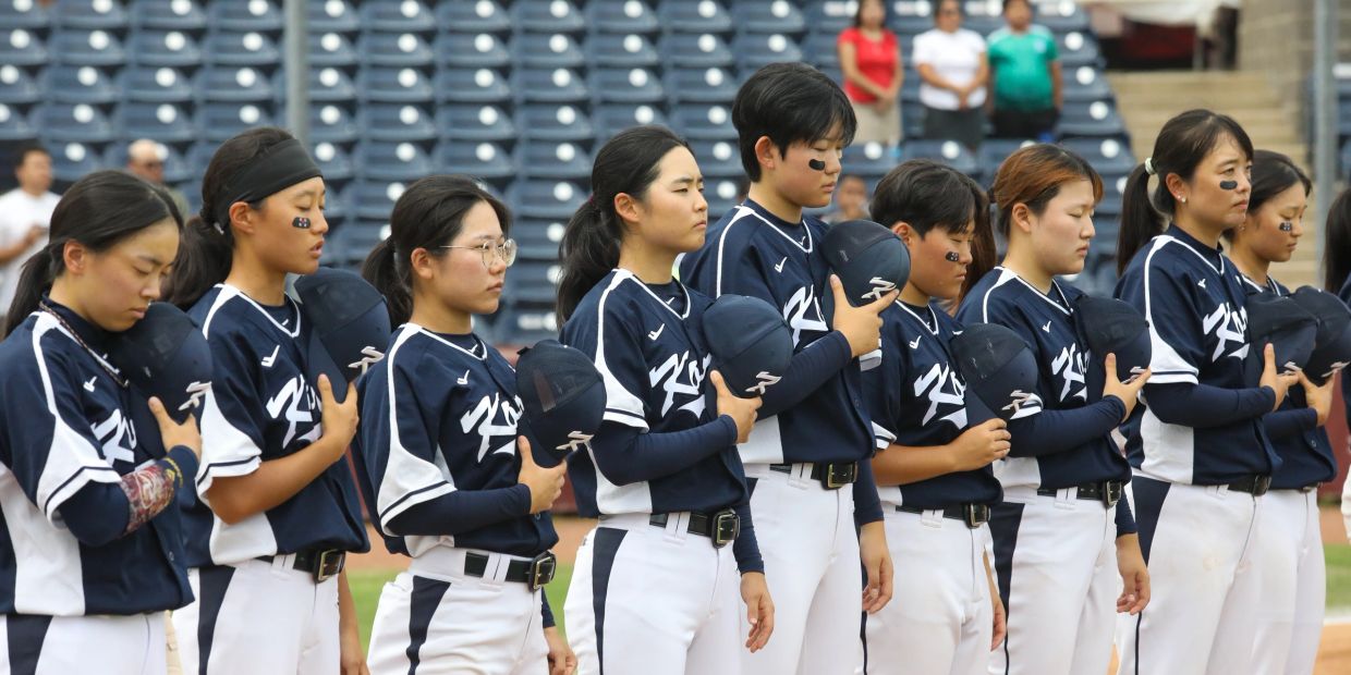 Female baseball team standing in line with hats over hearts.