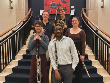 Five musicians stand together each holding either a french horn, flute, oboe, clarinet, or bassoon