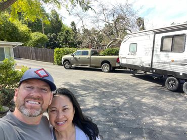 Happy couple taking selfie in driveway with a truck and camper trailer behind them.
