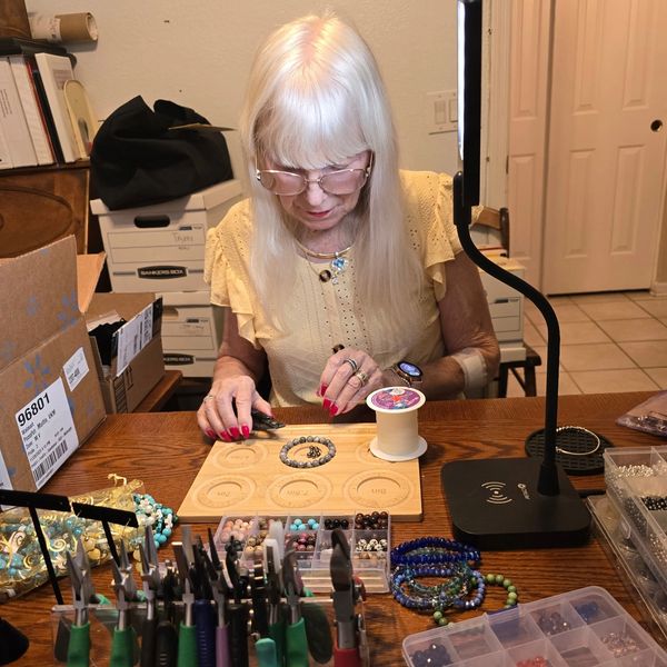 Elderly woman crafting beaded jewelry at a cluttered wooden table.