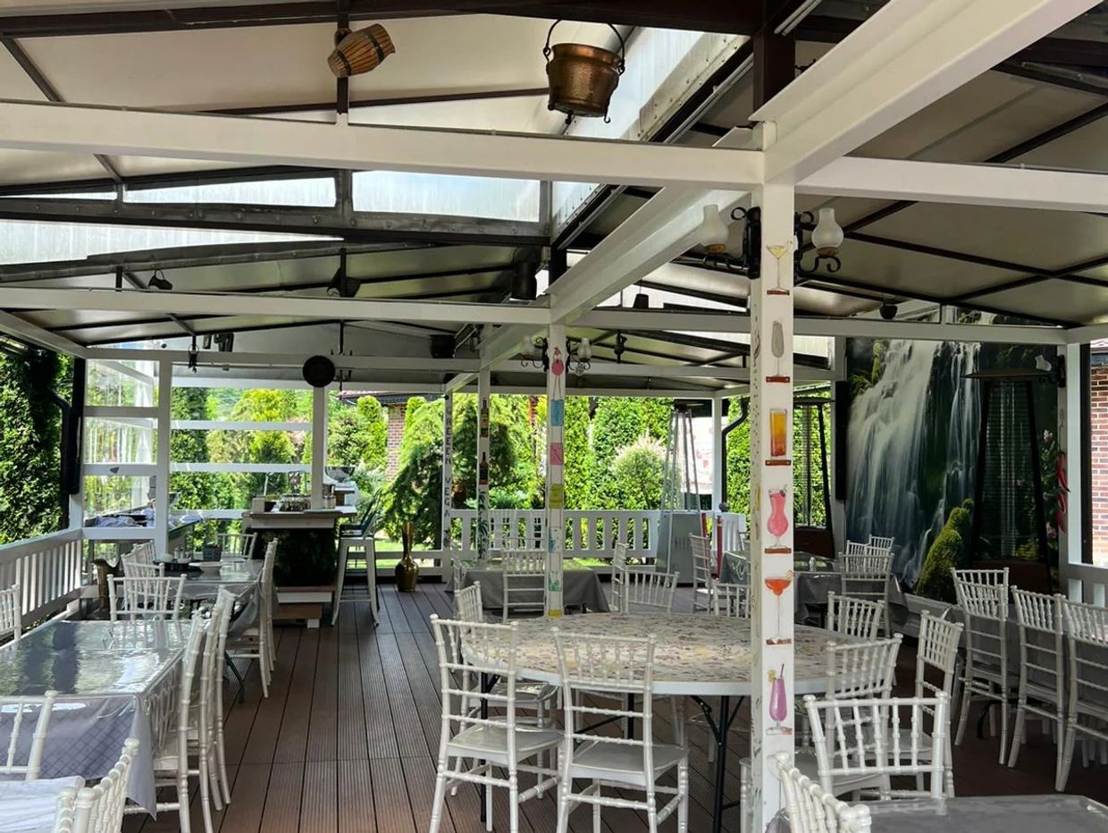 Covered outdoor dining area with white chairs and tables, surrounded by greenery.
