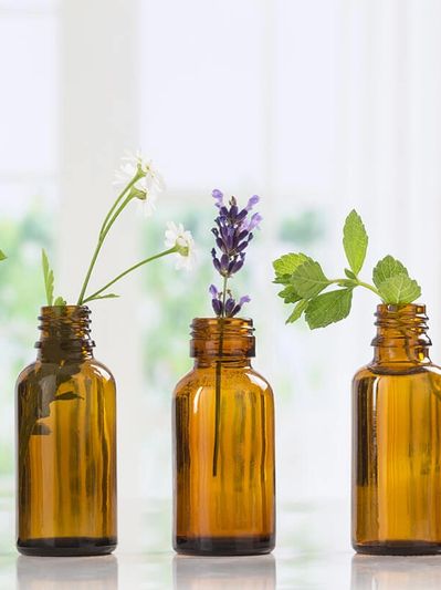 Five amber glass bottles each holding different fresh herbs and flowers on a white surface.