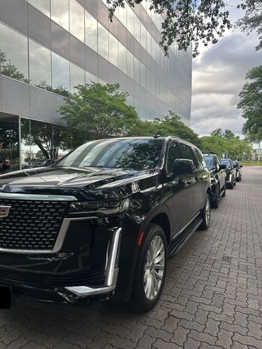 A lineup of black Cadillac SUVs parked along a sidewalk next to a modern glass building.