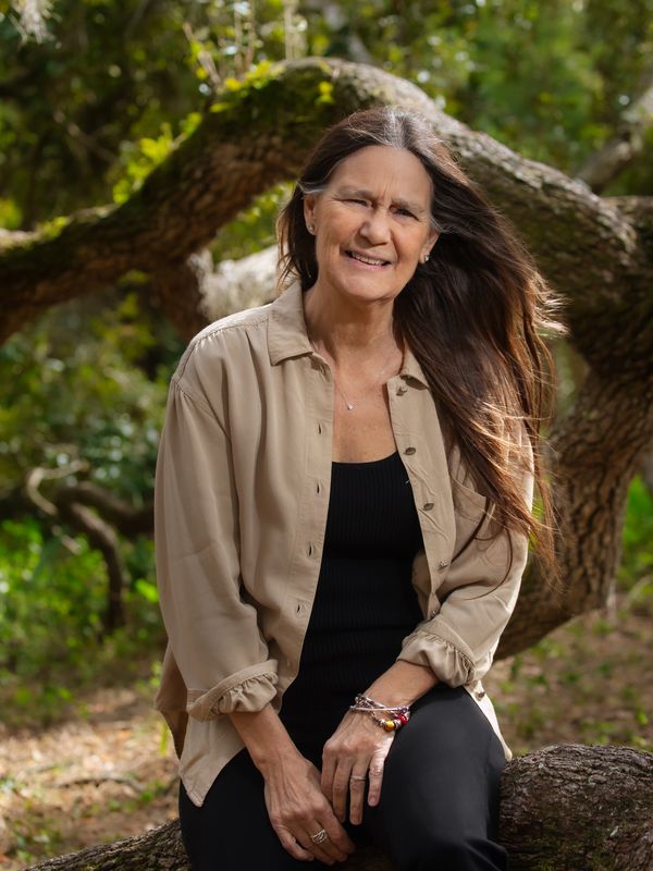 Smiling woman sitting outdoors with flowing hair and relaxed attire.