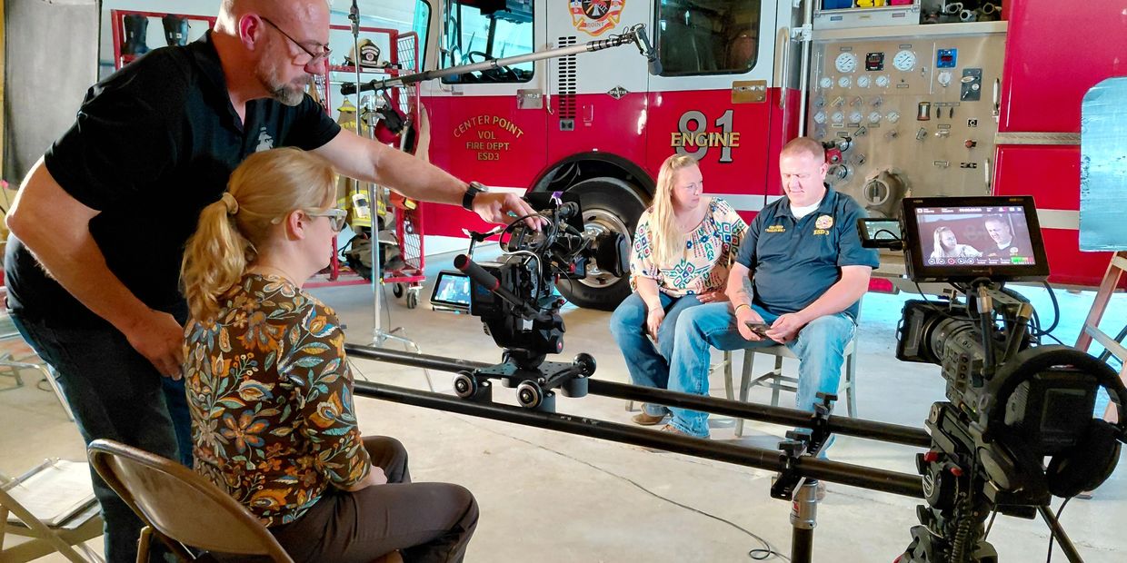 Film crew shoots an interview at a fire station with fire truck in background.