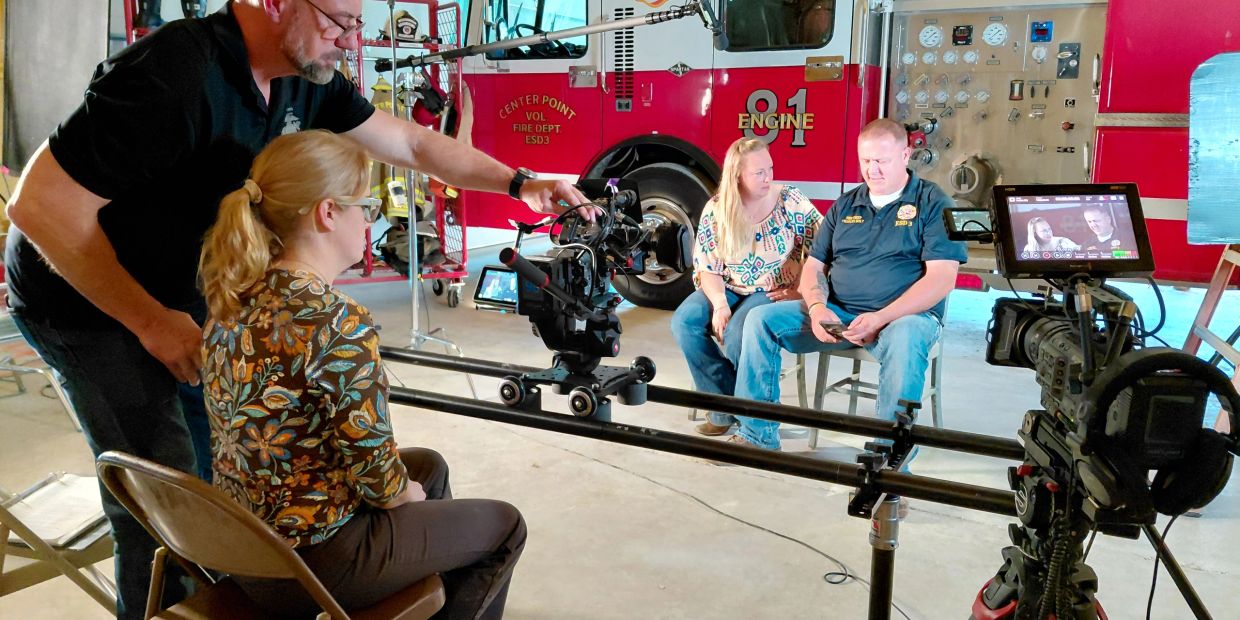 Film crew shoots an interview at a fire station with fire truck in background.